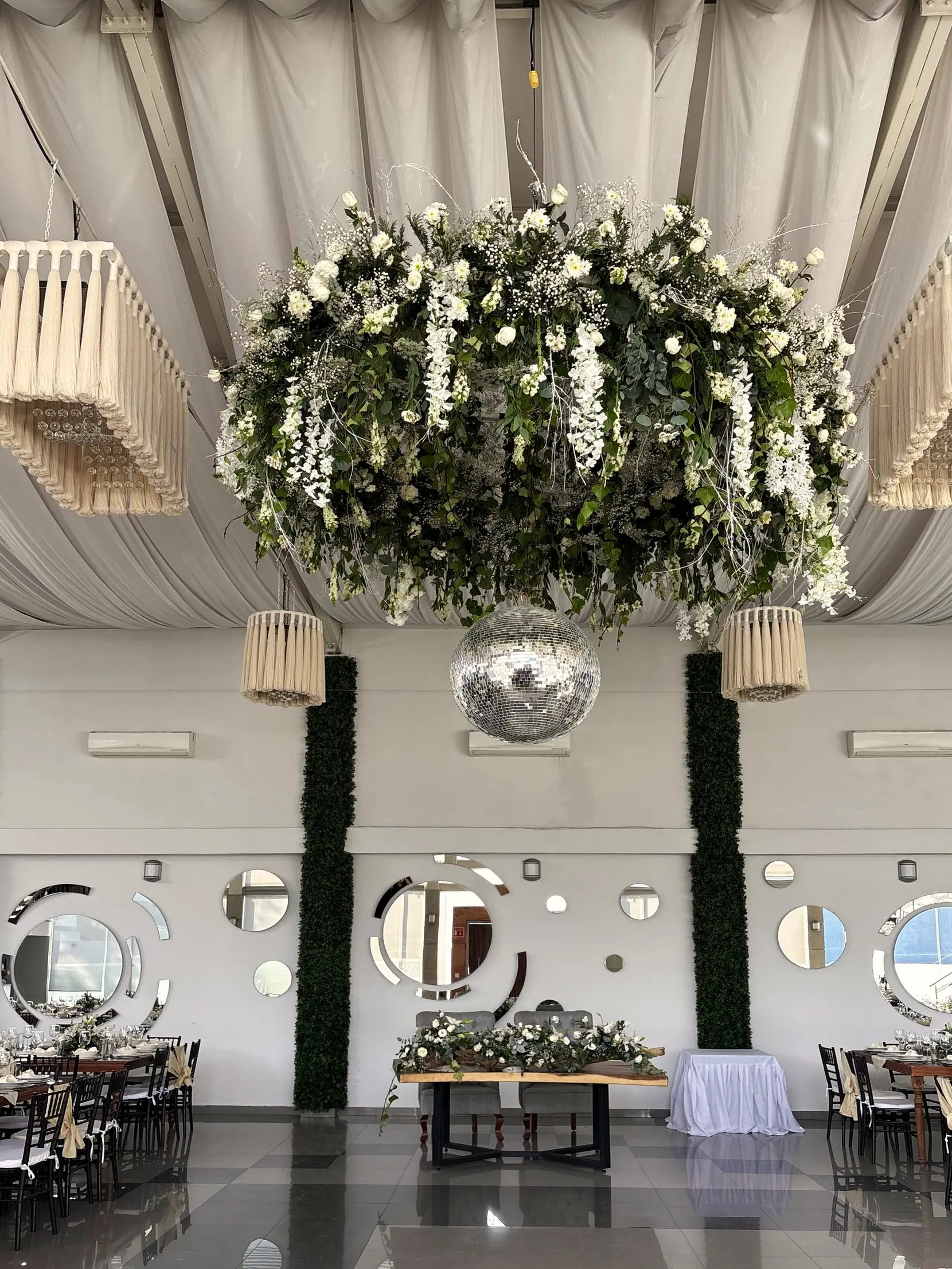 Corona de follaje y flores blancas bajo las lámparas de madera, con la mesa de novios al fondo