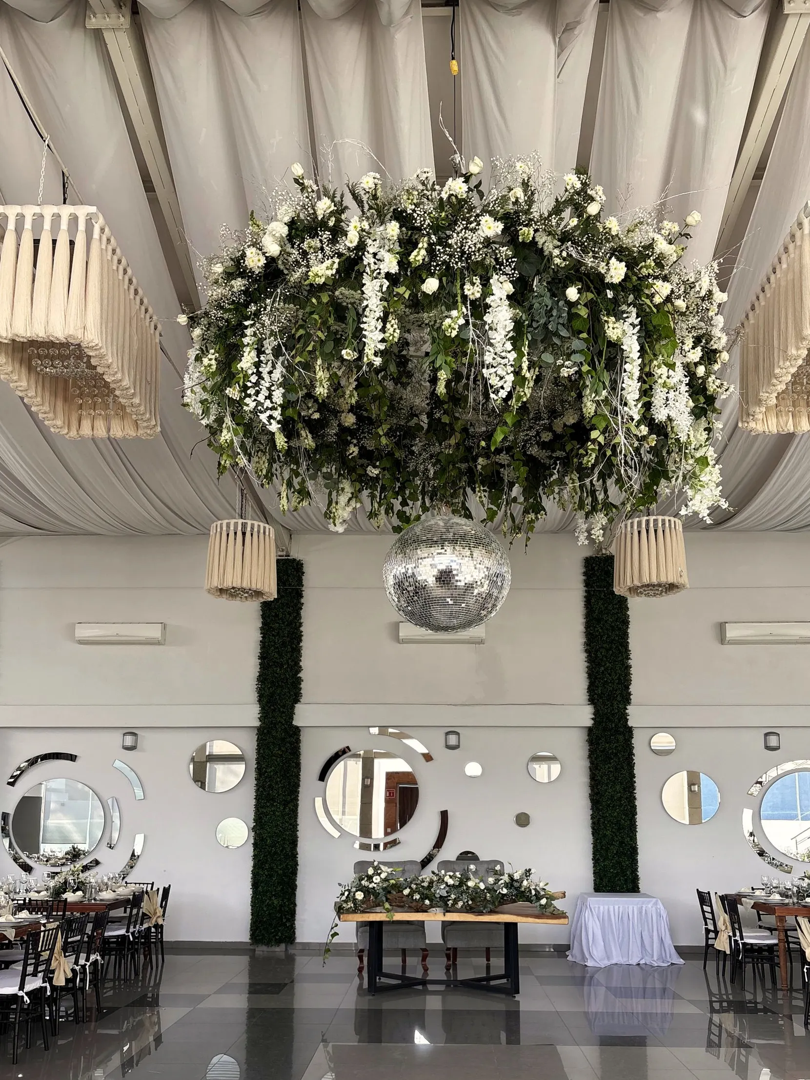 Corona de follaje y flores blancas bajo las lámparas de madera, con la mesa de novios al fondo